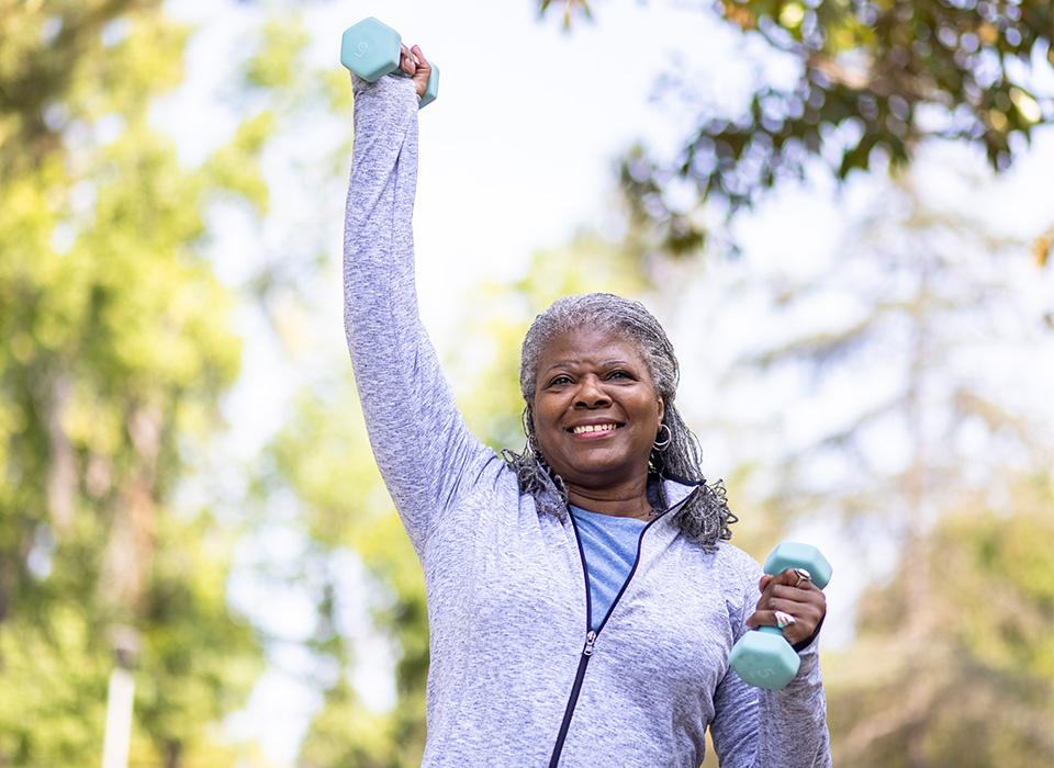 gray-haired woman exercising outside with light dumbbells, with one arm raised above her head